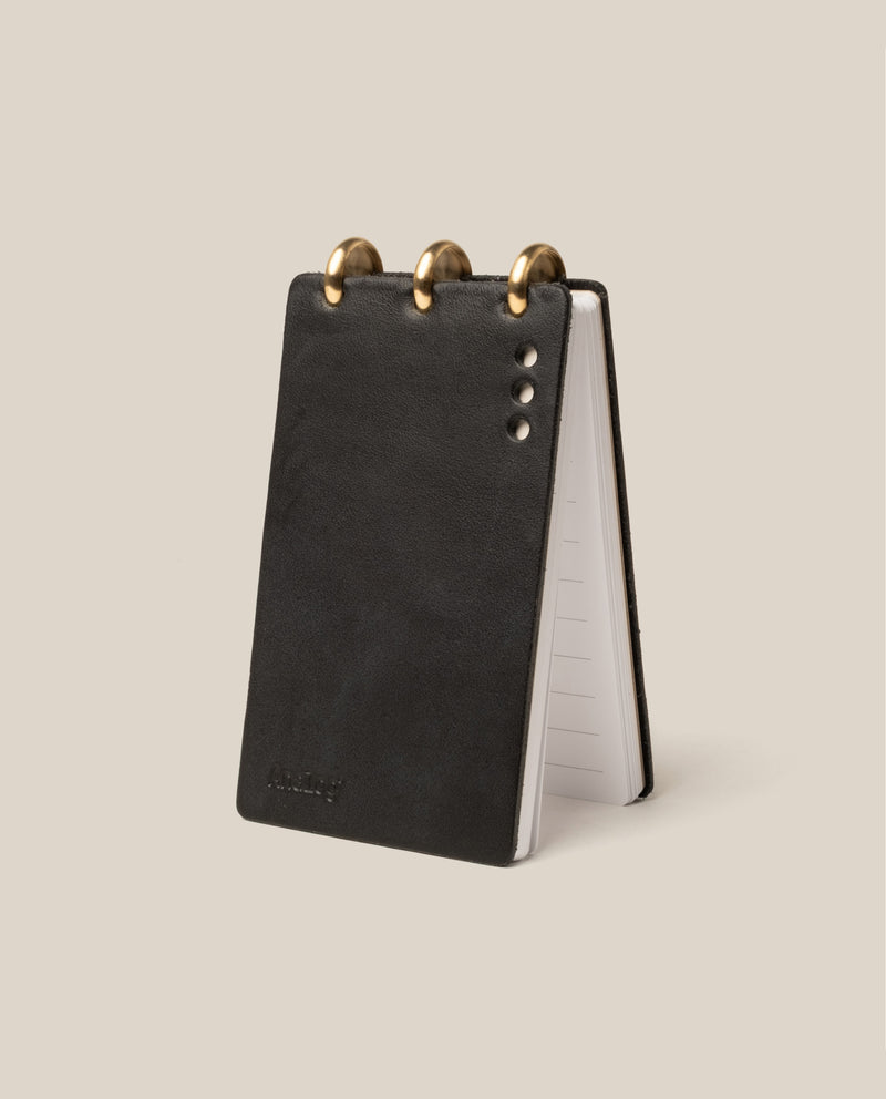 A small black leather notepad with three brass rings at the top, standing upright and slightly open, revealing lined pages inside. The cover has three metallic rivets and a subtle embossed logo.