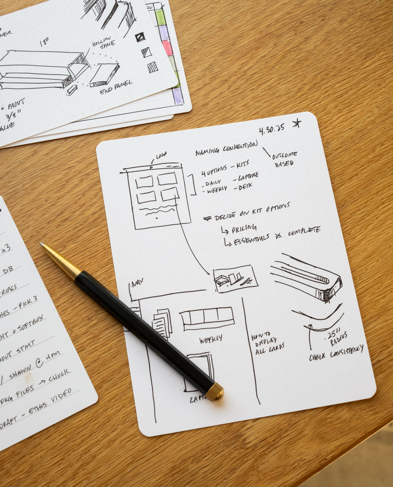 A black pen rests on a notepad with hand-drawn project diagrams, alongside Analog Dot Grid Cards - Large (12-Pack) and 100lb card stock, all arranged on a wooden desk scattered with notes and papers.