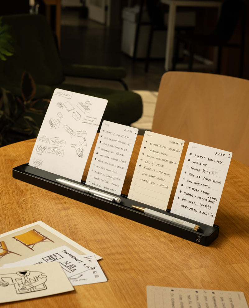 A desk organizer displays several notecards with handwritten notes and sketches, along with two pens. The organizer sits on a wooden table in a cozy, well-lit room with chairs and plants in the background.