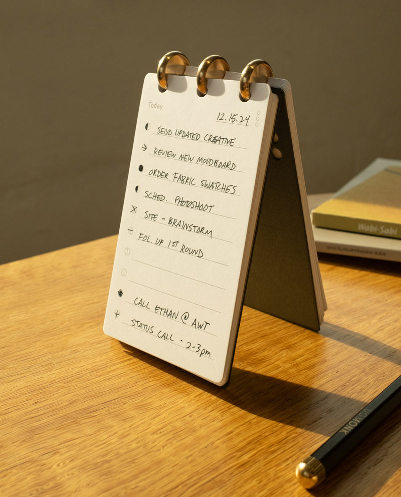 A Discbound Analog flip desk calendar, filled with handwritten to-do lists, stands on a wooden table beside a pencil, several closed books, and a refillable note card stack—an essential part of any analog task system—illuminated by warm sunlight.