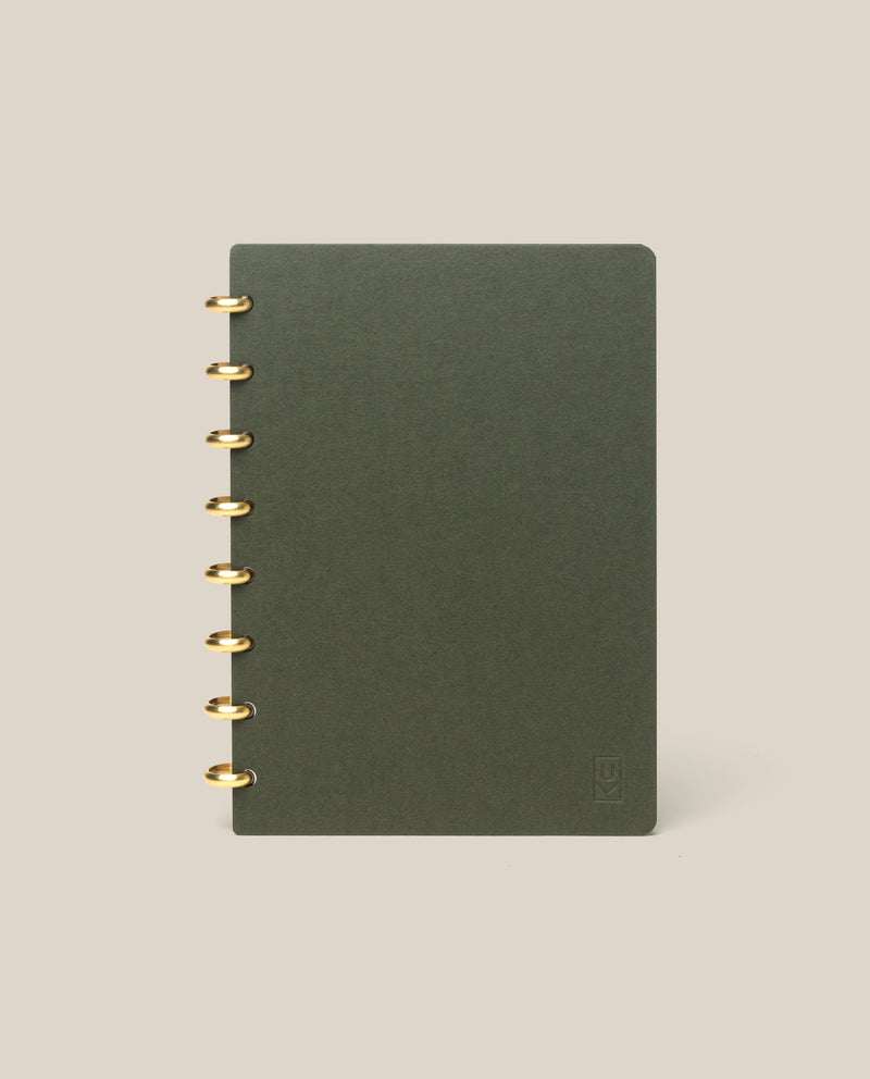 A Discbound Journal (Standard 6"x 8.5") in dark green with brass discs on the left stands upright against a plain beige background.