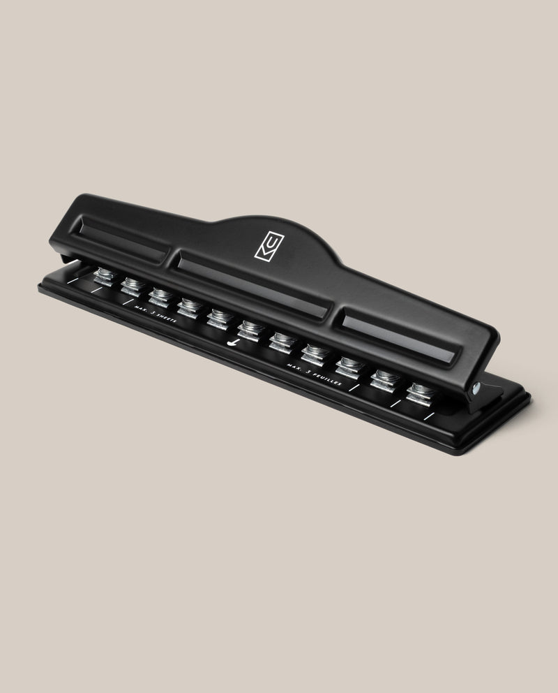 A black metal Discbound Punch with a lever handle is displayed on a light beige background. It features measurement markings and metal cutters for punching holes compatible with the Ugmonk Discbound Journal or any discbound system.
