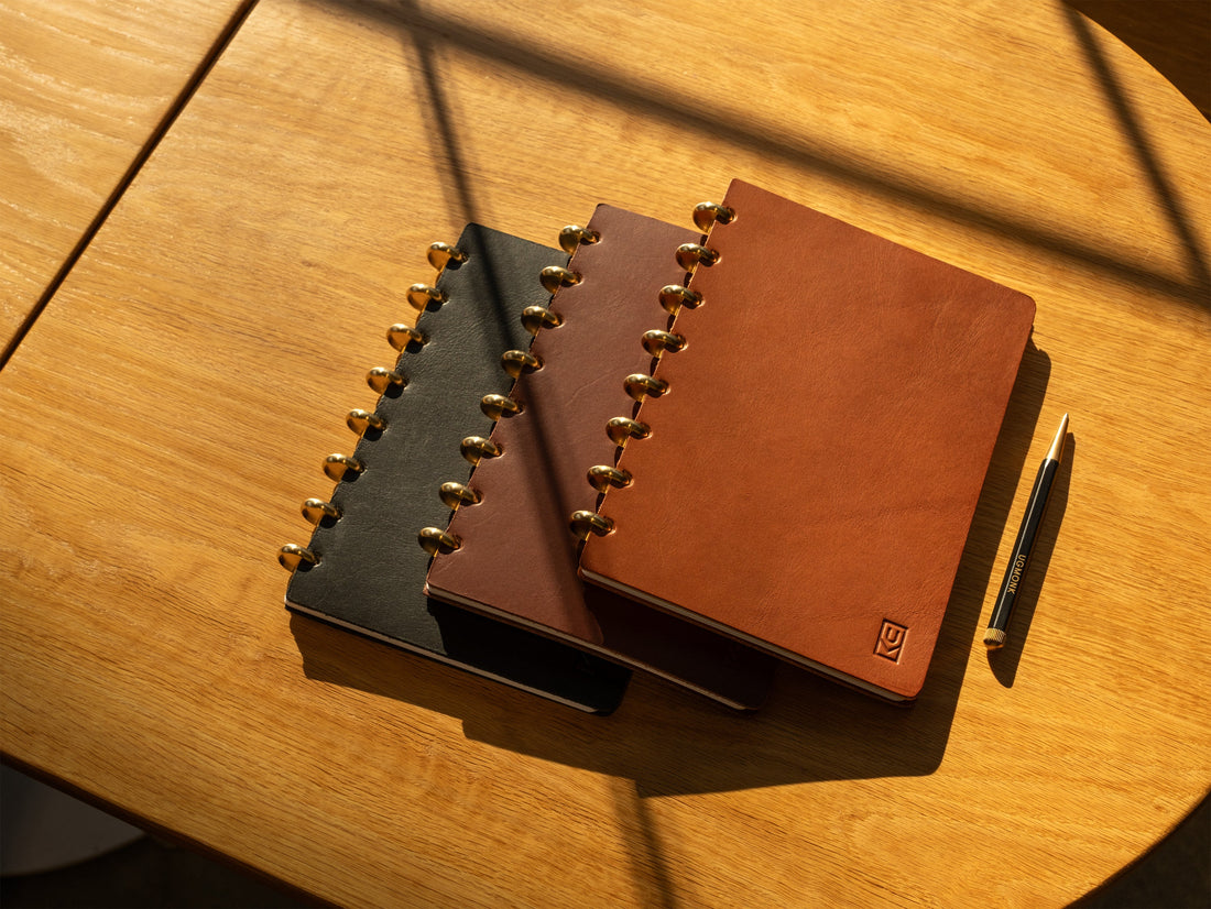 Three leather-bound notebooks with gold rings and a pen rest on a wooden table in warm sunlight, casting distinct shadows across the surface.