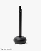 A sleek desk accessory, the HMM Paperweight Pen Stand (Black) features a round, weighted aluminum base and a vertical slot for upright pen storage. Plain white background. Text at bottom: Pen sold separately.