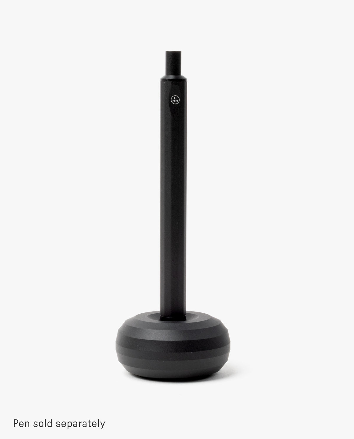 A sleek desk accessory, the HMM Paperweight Pen Stand (Black) features a round, weighted aluminum base and a vertical slot for upright pen storage. Plain white background. Text at bottom: Pen sold separately.