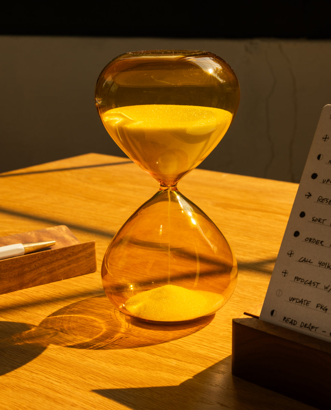 The 25 Minute Hourglass (Amber), a hand-blown glass timer with yellow sand, sits on a wooden desk in warm sunlight beside a pen and notepad.