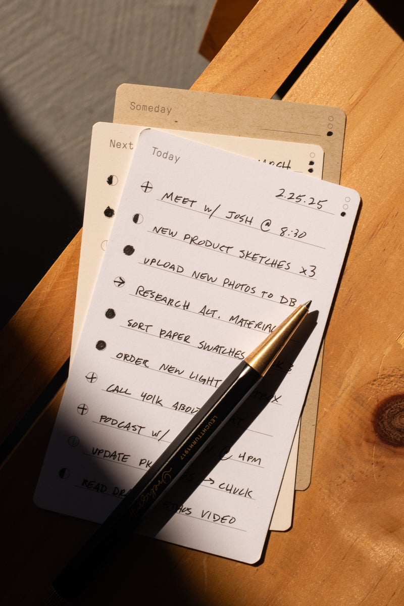 The Analog Daily Focus Kit (Walnut) displays a handwritten to-do list with tasks like meeting with Josh, product sketches, and uploading photos on a wooden surface beside a black pen and additional task cards for organized productivity.
