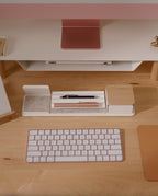 A neatly organized wooden desk features a white and pink keyboard, a matching monitor, pen holder, pen, notepad, and a small wooden box, all arranged tidily atop the Gather 1x4 Base Plate (White), which is powder-coated steel and designed to securely hold magnetic Gather modules.