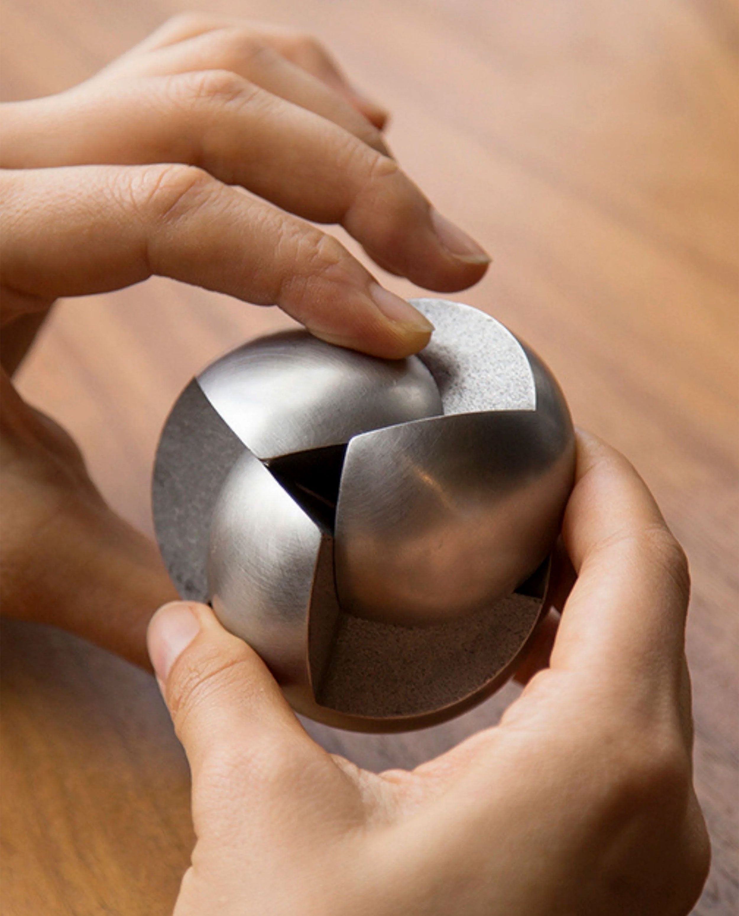 A person’s hands are holding and manipulating the Craighill Venn Puzzle (Stainless Steel), with its interlocking curved pieces forming a sphere, against a wooden surface background.