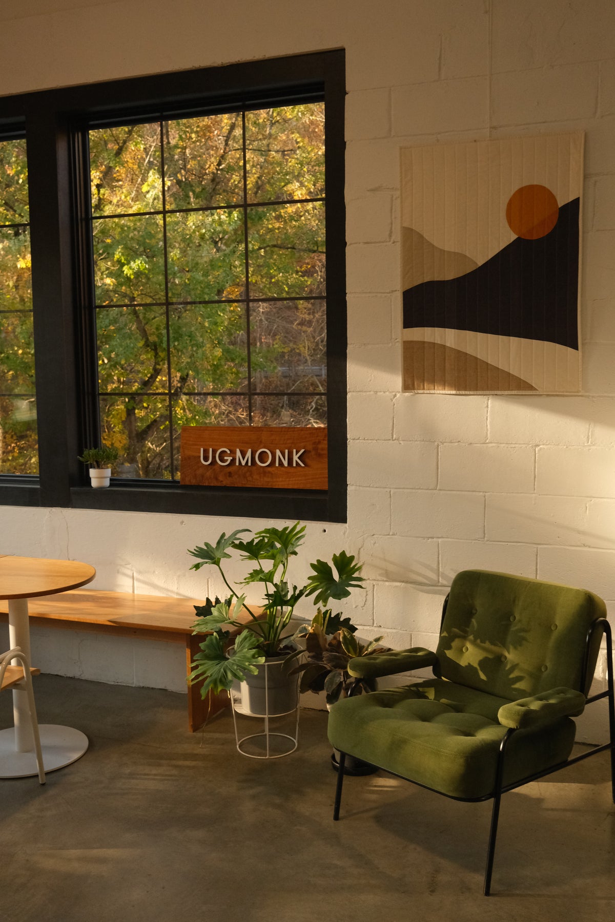 Sunlit modern room with a green armchair, small round table, plants, and wall art. Large windows reveal trees with green leaves outside. A wooden sign reading UGMONK sits on the bench below the window.