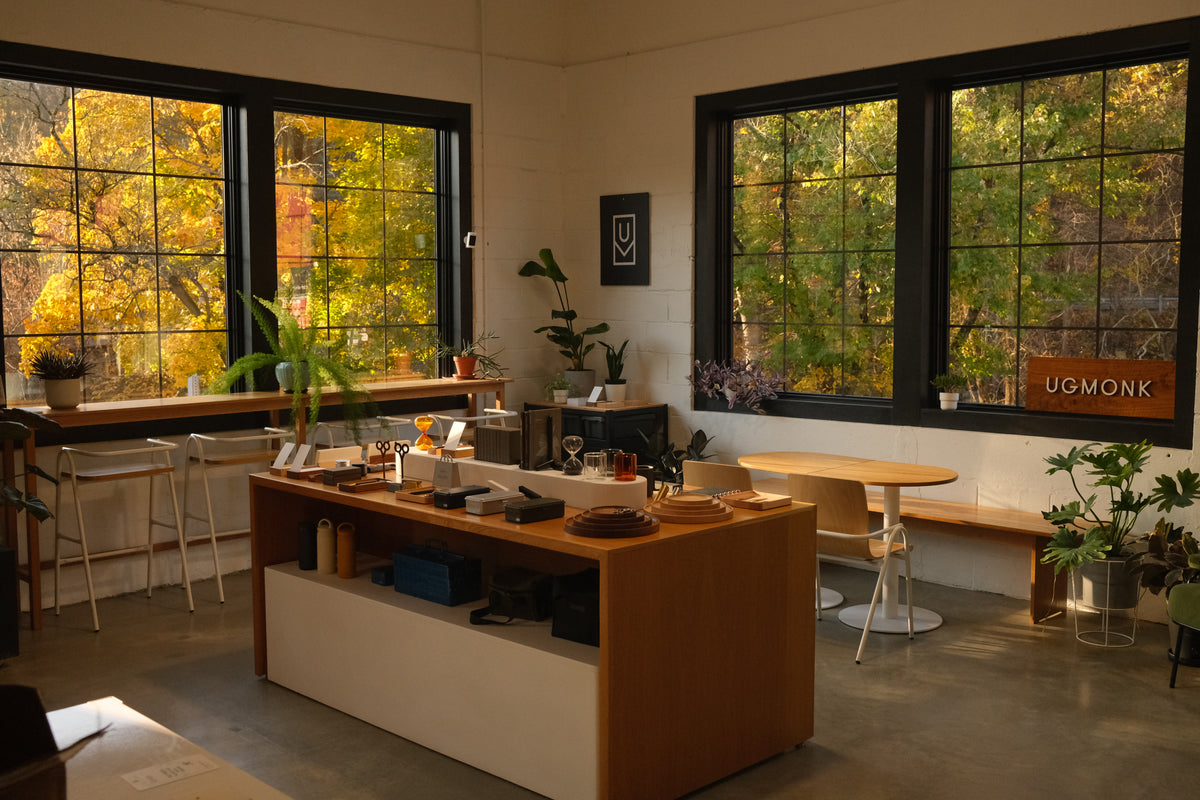 A cozy, sunlit store interior with wooden furniture, plants, neatly arranged items on a central counter, and large windows showcasing autumn trees outside.