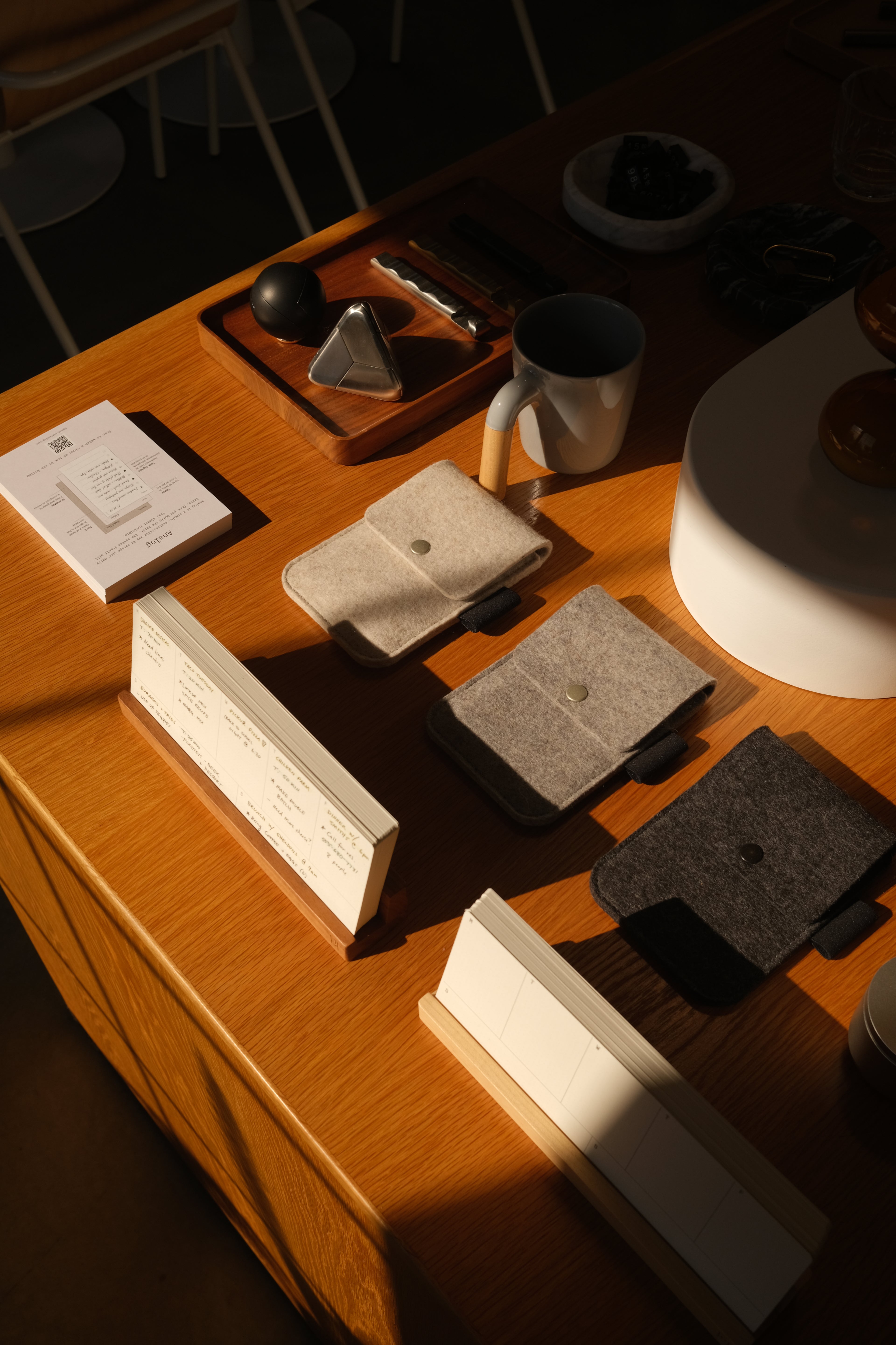 A wooden table displays felt wallets in gray and beige, a ceramic mug, a tray with utensils, paper notepads, and a menu, all lit by warm sunlight and deep shadows.