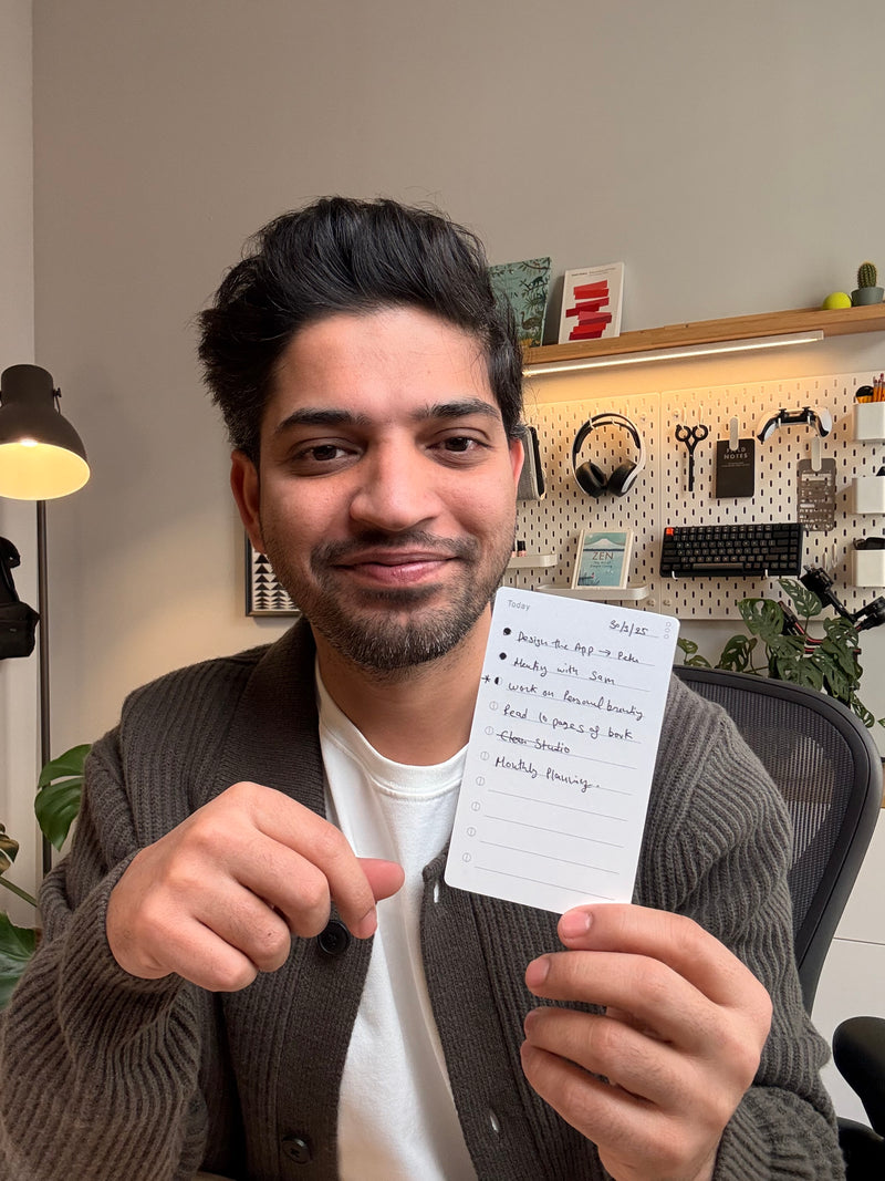 A person smiling and holding up a handwritten to-do list in an office with a plant, desk lamp, and organized tools on the wall behind them.