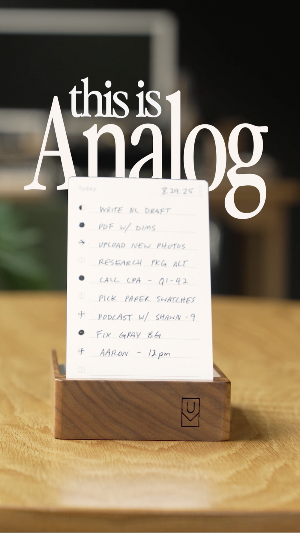 A handwritten to-do list sits in a wooden holder on a desk. Large text above reads “this is Analog.” The blurred background shows a computer screen and other office items.