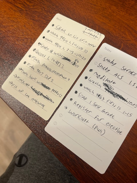 Two handwritten to-do lists on small white cards rest on a wooden table. Tasks include workout, register for elective, finish paperwork, and several items are crossed off, indicating completion. Part of a gray fabric is visible below.