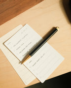 A notepad with a to-do list and The Ugmonk Pen (Leuchtturm1917 Drehgriffel - Black) placed diagonally on top, resting on a wooden desk. Tasks include The Compass Collection, Bigger Item, Copy, and Launch Party Set Up.