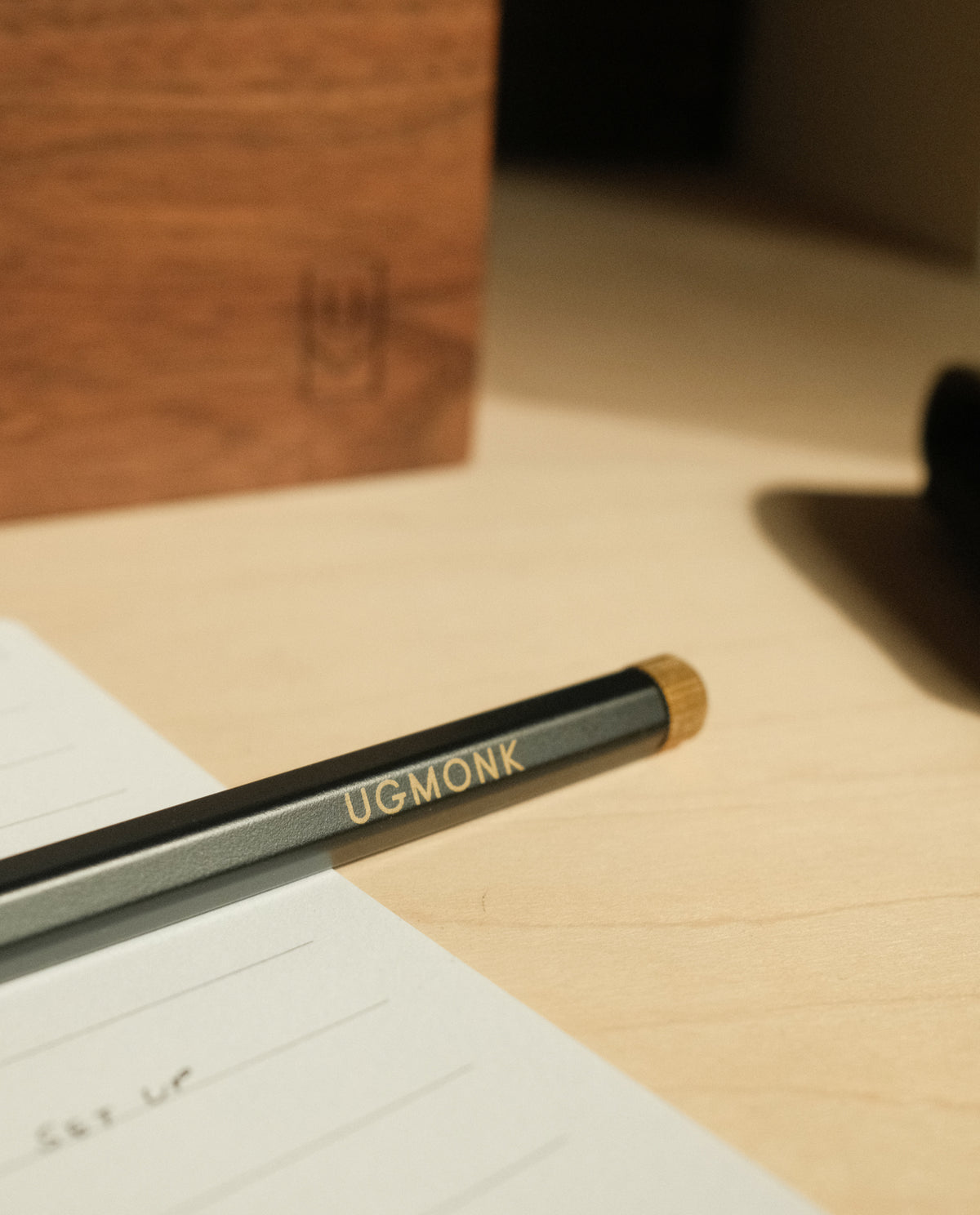 A close-up of a pencil engraved with UGMONK rests on a lined notebook atop a light wooden desk, with The Ugmonk Pen (Leuchtturm1917 Drehgriffel - Black) and a wooden box bearing a subtle logo softly blurred in the background.