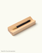 The Ugmonk Pen (Leuchtturm1917 Drehgriffel - Black) sits on a light wooden pen tray against a plain white background. Text below says, “Wood Pen Tray sold separately.”.