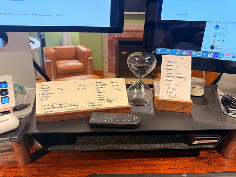 Two monitors on a desk with handwritten notes, an hourglass, and a remote control in front. A brown chair and fireplace are visible in the background.