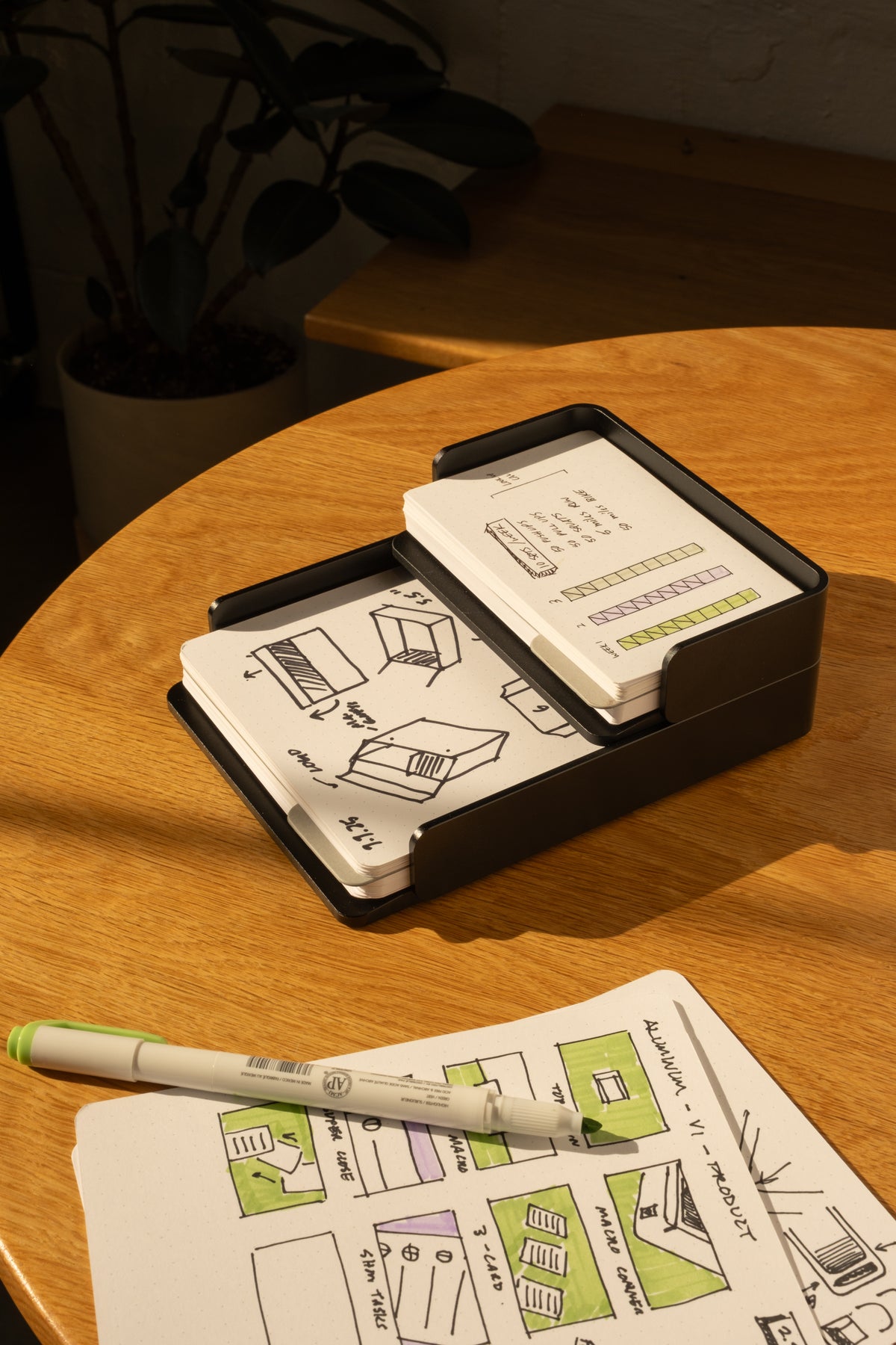 The Analog Capture Kit - Large (Black Aluminum) features illustrated dot grid cards in a sleek black holder on a wooden table, paired with a pen and open notebook. A potted plant adds to the creative workspace.
