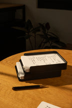 The Analog Capture Kit - 2 Large (Black Aluminum) sits on a round wooden table with dot grid cards and a black pen, bathed in sunlight with warm shadows and a blurred plant in the background.