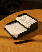 The Complete Analog Collection (Black Aluminum) two-tier card organizer sits on a wooden table holding index cards with diagrams and handwritten notes. Nearby is a black and gold pen, with a blurred plant in the background.