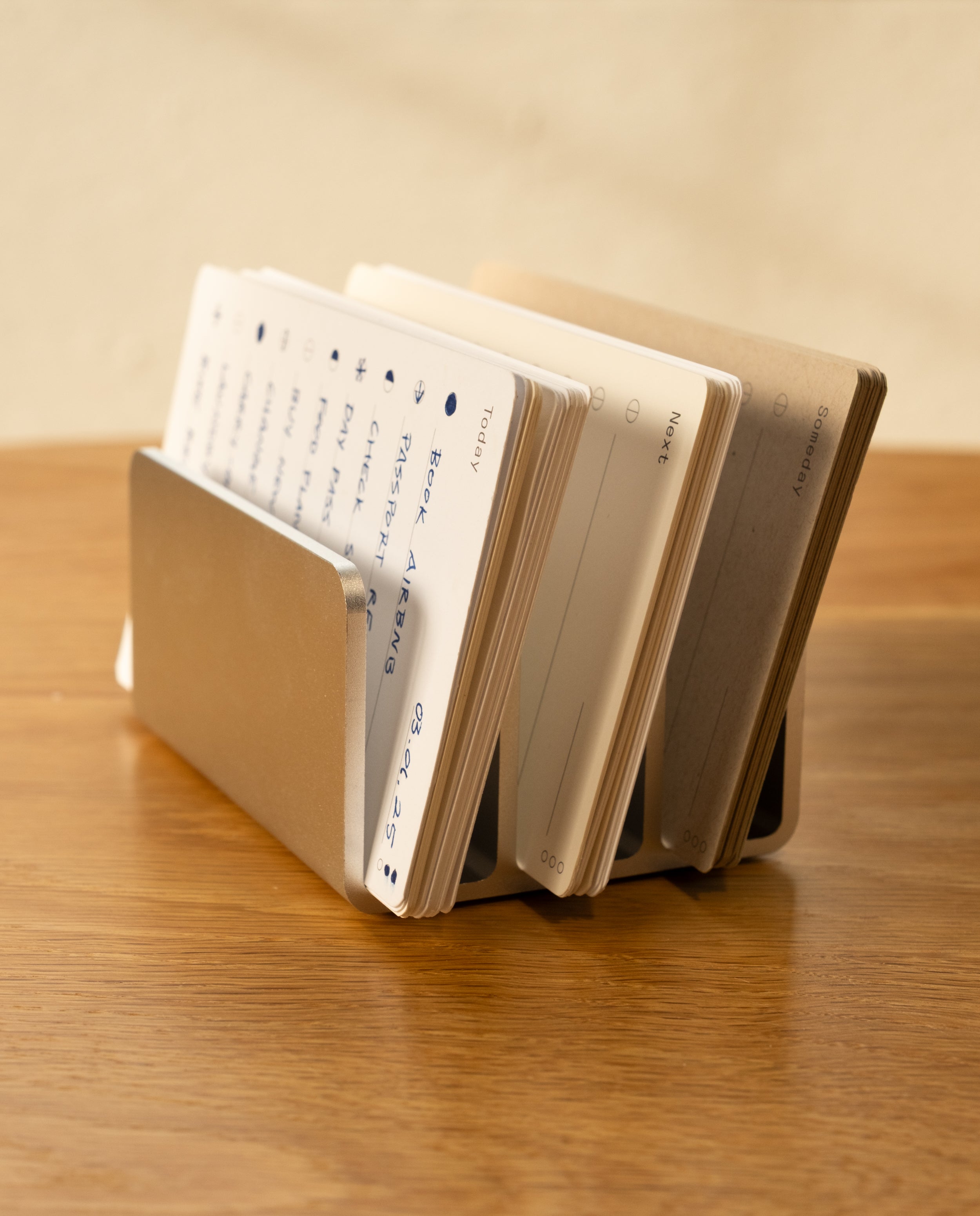 The Analog Card Organizer (Silver Aluminum) holds index cards with handwritten notes, fanned out and labeled by day, resting on a wooden surface.