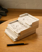 The Analog Capture Kit - 2 Large (Silver Aluminum) sits on a wooden desk, holding illustrated instruction cards. A black and gold pen is nearby, with part of a gray item and a light-colored wall in the background.