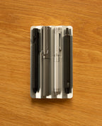 Four pens in black, gray, silver, and dark gray are neatly arranged side by side in the Analog Multi Pen Tray (Silver Aluminum) on a wooden surface.