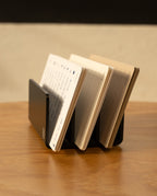 The Analog Card Organizer in black aluminum displays several open notebooks with lined and blank pages on a wooden surface against a light-colored wall.