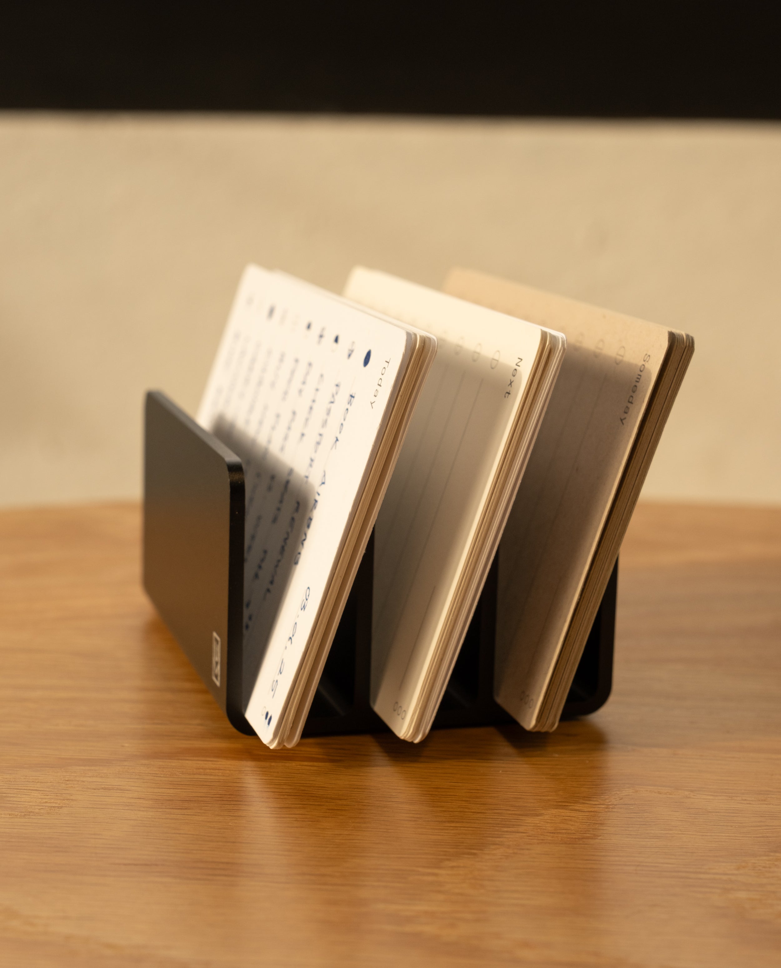 The Analog Card Organizer in black aluminum displays several open notebooks with lined and blank pages on a wooden surface against a light-colored wall.