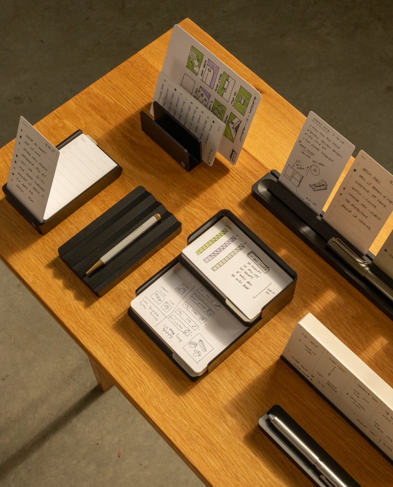 A wooden table displays The Complete Analog Collection (Black Aluminum), organizing notepads, pens, and sketches. The well-lit scene features a plain gray floor in the background.