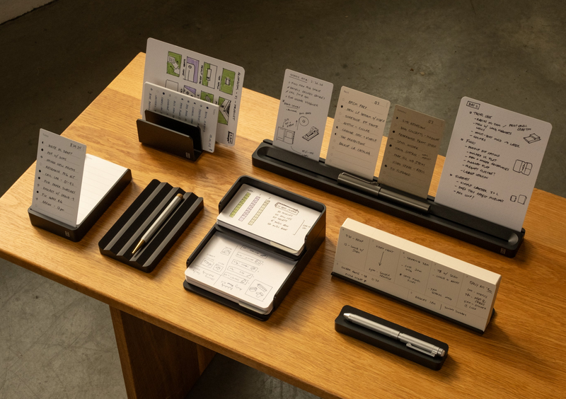 A wooden desk displays multiple black organizers holding notepads, index cards, and pens, each with handwritten notes, sketches, and diagrams. The background is an empty, gray concrete floor.