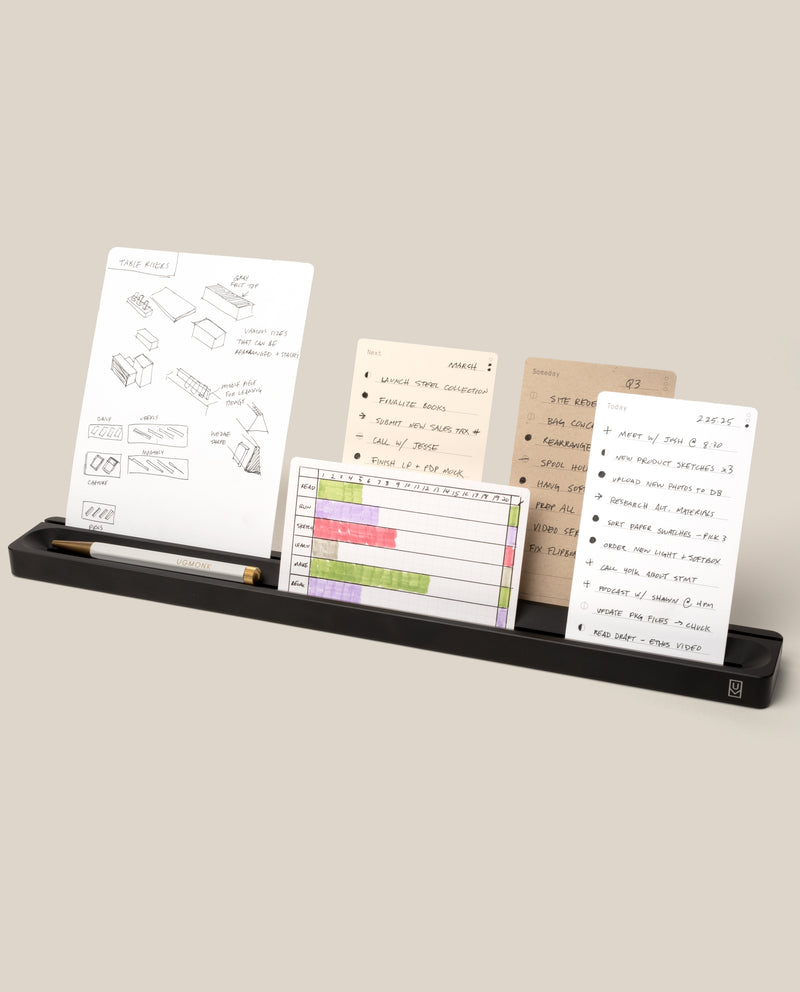 The Analog Card Bar (Large 16" – Black) is a sleek black desk organizer, styled like a solid aluminum card holder and desktop pen tray, that holds a pen and four notecards displaying sketches, lists, handwritten notes, and a color-coded chart against a beige background.