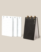 The Leather Discbound Analog + 3 Refills (Black), featuring full-grain leather covers, is shown upright on a neutral background next to three stacks of daily planner refill pages.