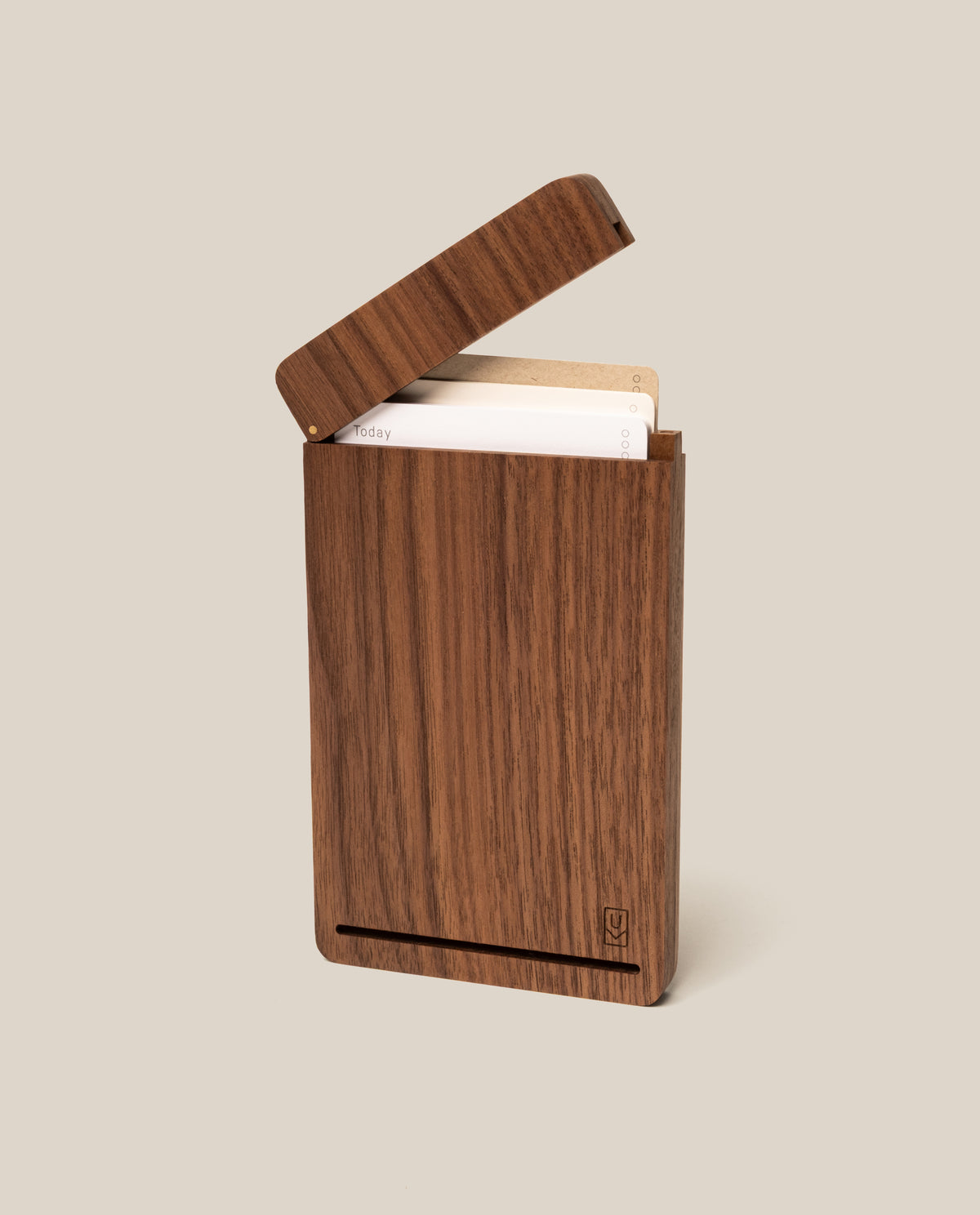 A walnut Analog Travel Case with a hinged lid, shown partially open to reveal cards inside. The case features a minimal design with a small logo engraved at the bottom right corner and is equipped with a secure magnetic closure. The background is plain beige.