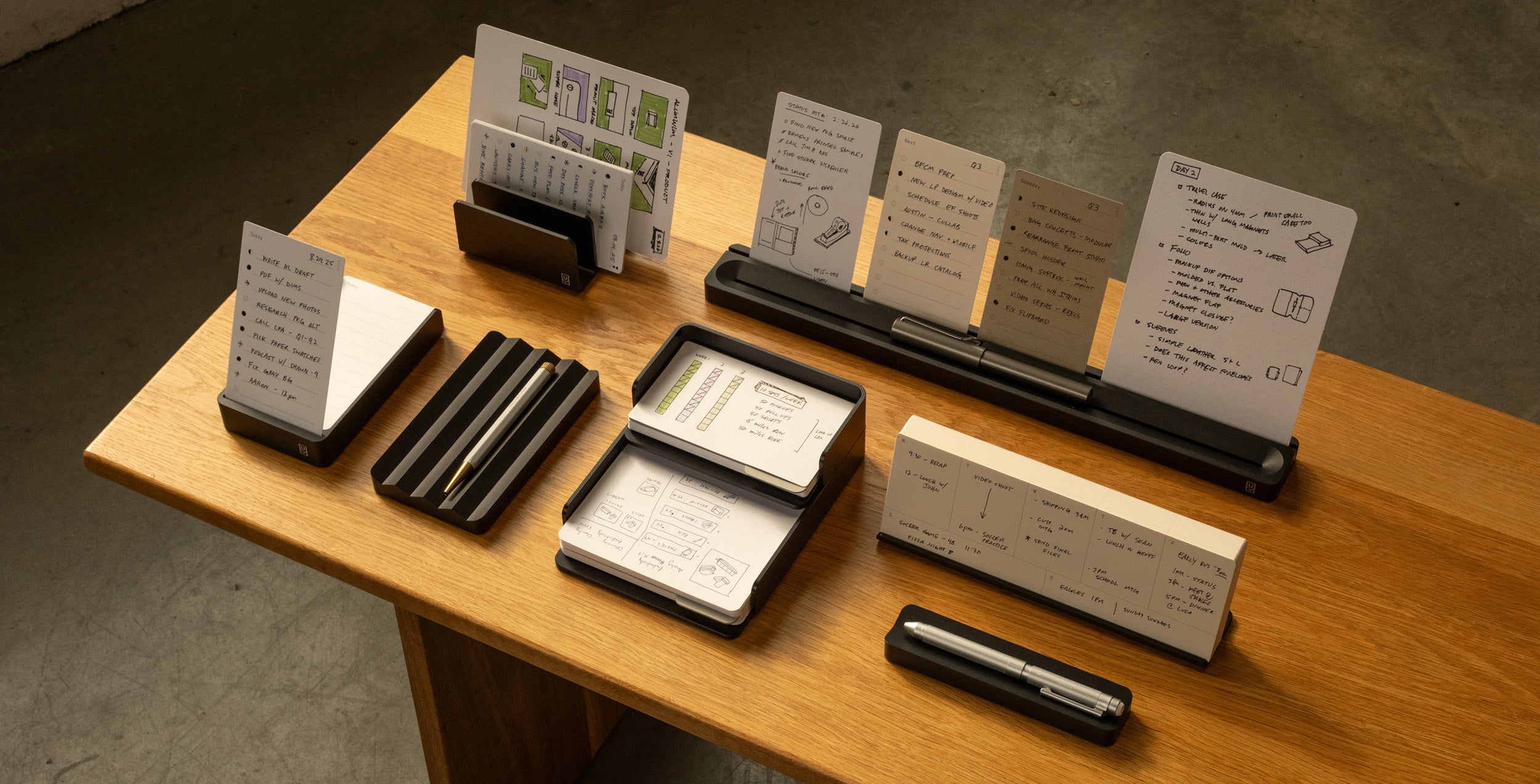 A wooden desk displays various black desk organizers holding notecards with handwritten notes and sketches, a silver pen, and a stack of cards, all arranged neatly under warm lighting.