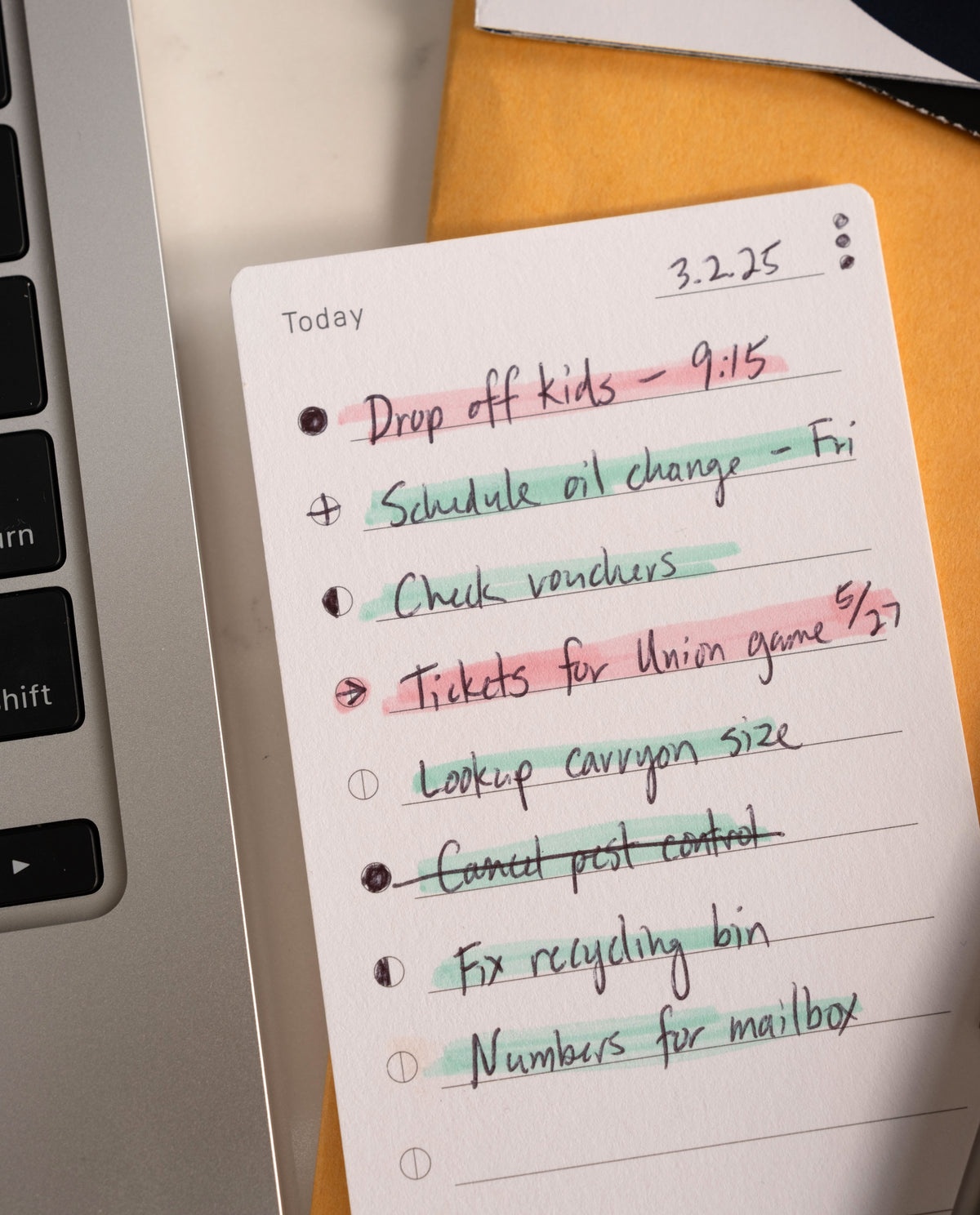 A set of Analog Task Cards - Small, featuring handwritten tasks like dropping off kids and scheduling an oil change—some checked off or crossed out in various colors—lies beside a laptop, exemplifying a straightforward analog system in use.