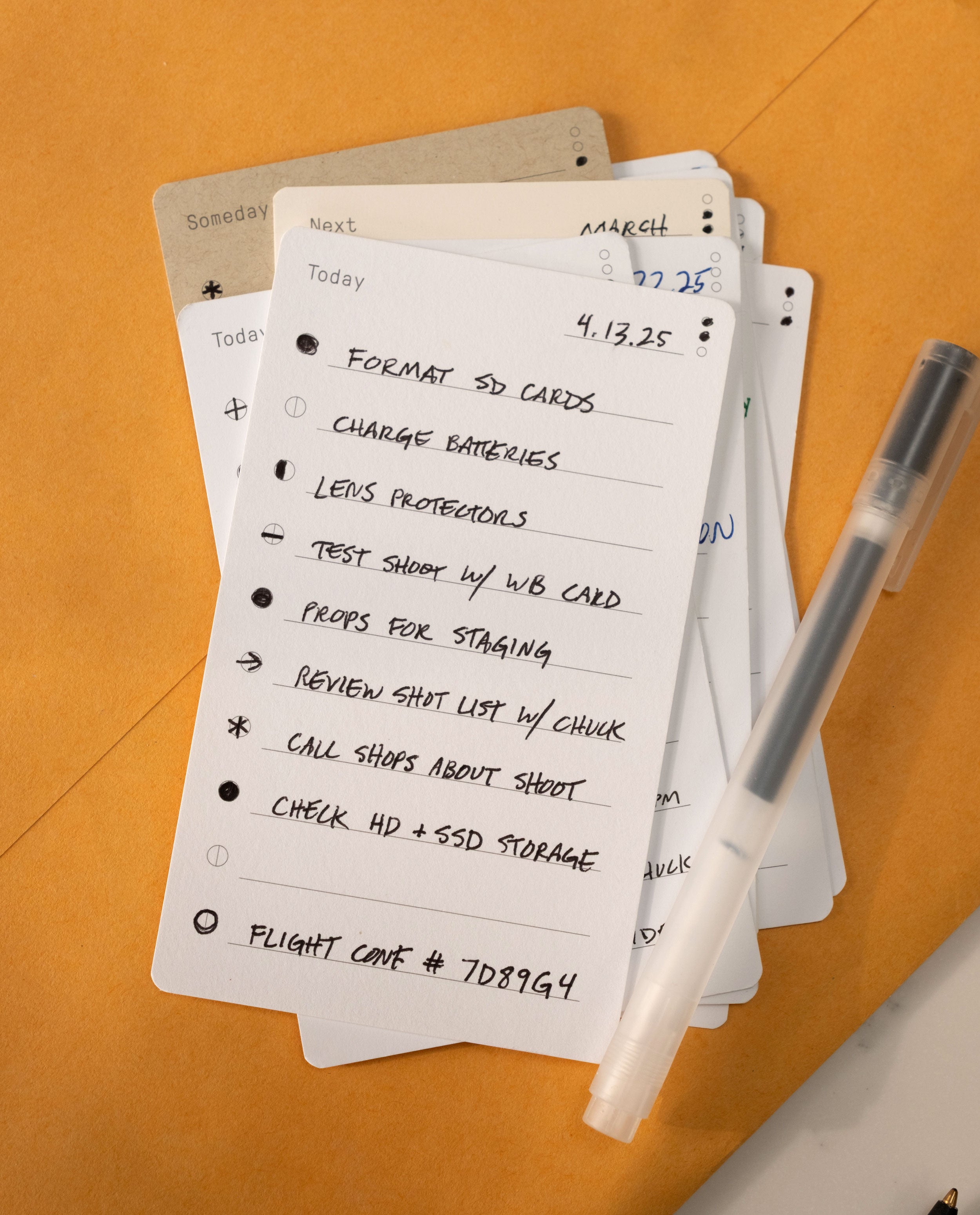 A handwritten to-do list for a photoshoot, created on Analog Task Cards - Small, sits atop other productivity cards with a pen nearby. Tasks include formatting SD cards, charging batteries, and checking equipment—demonstrating an effective analog system for staying organized. A flight confirmation code is noted at the bottom.