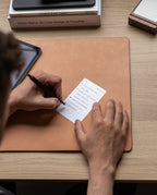 Someone writes a to-do list on an Analog Annual Refill card while sitting at a desk with a leather mat, tablet, and stacked books, including Dieter Rams: As Little Design as Possible.