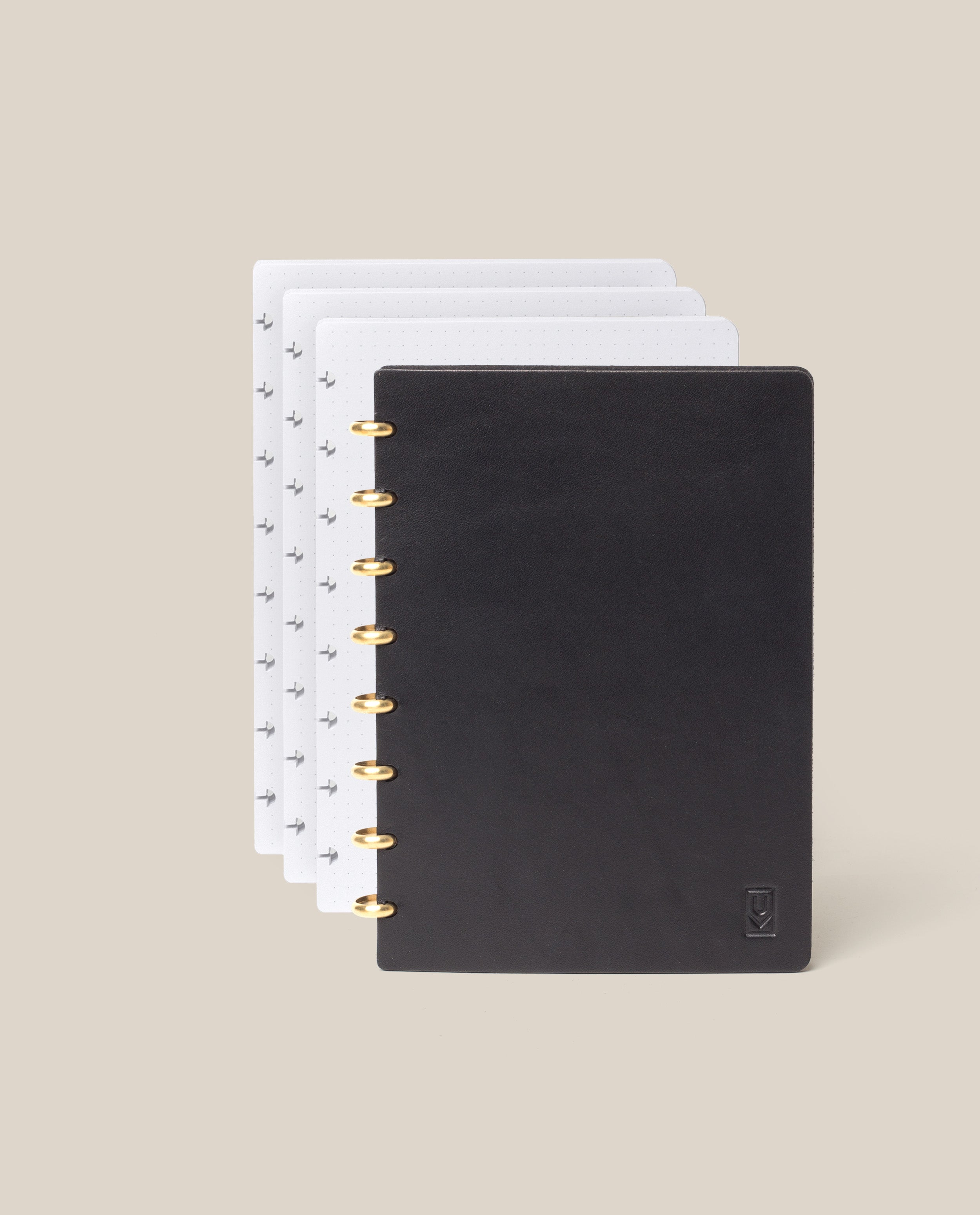 The Discbound Heirloom Journal Bundle (Black Leather - Standard 6" x 8.5" + 3 Refills) is open with gold rings, showing dotted and patterned pages fanned out behind the cover on a light beige background.