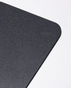 A close-up of the rounded corner of a textured, rectangular black object—specifically, the Gather 2x2 Base Plate (Black), crafted from powder coated steel and designed to support magnetic Gather modules—set against a plain white background.