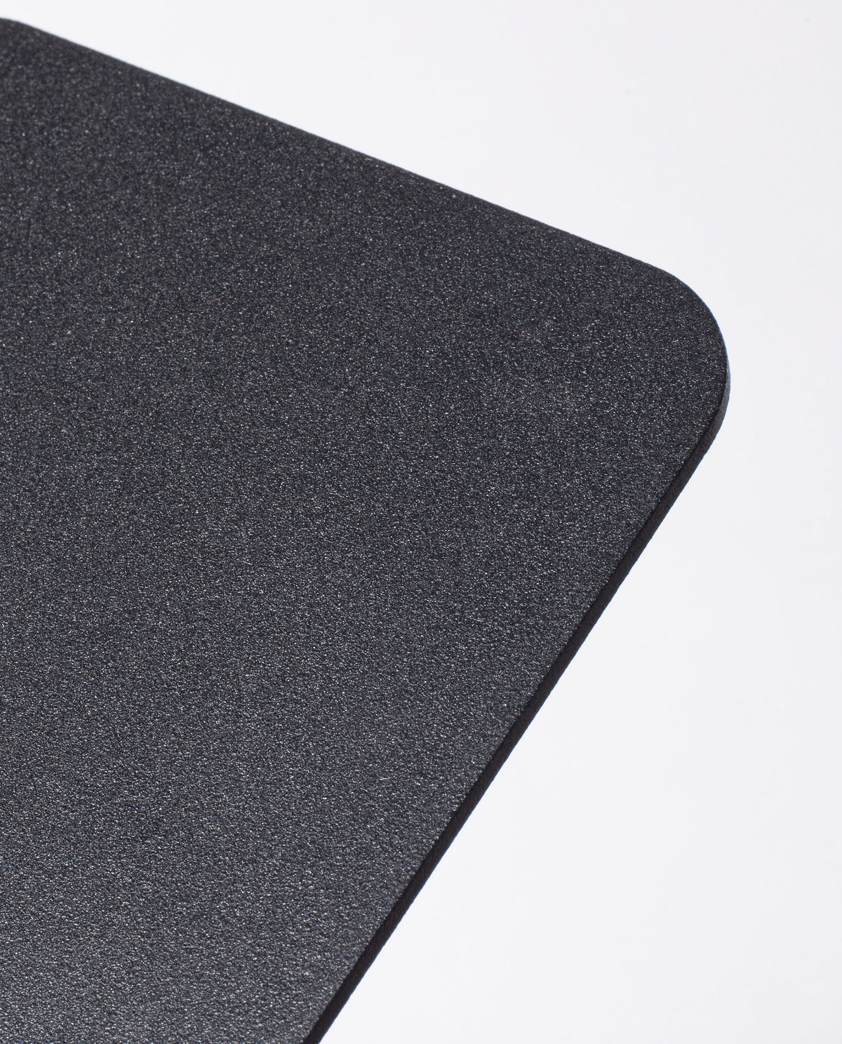 A close-up of the rounded corner of a textured, rectangular black object—specifically, the Gather 2x2 Base Plate (Black), crafted from powder coated steel and designed to support magnetic Gather modules—set against a plain white background.
