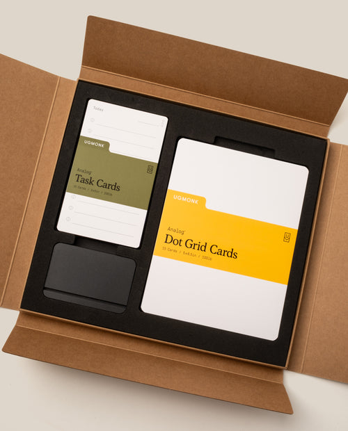 Open brown box displaying the Analog Starter Kit (Black) by Ugmonk, featuring Task Cards, Dot Grid Cards, and a black cardholder set in custom foam compartments—an elegant paper card system perfectly complemented by the optional aluminum card stand.