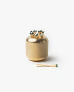The Craighill Match Striker (Brass) holds strike-anywhere matches upright in its modern, minimalist solid brass container with a textured surface. Two matches rest beside it on a white background.