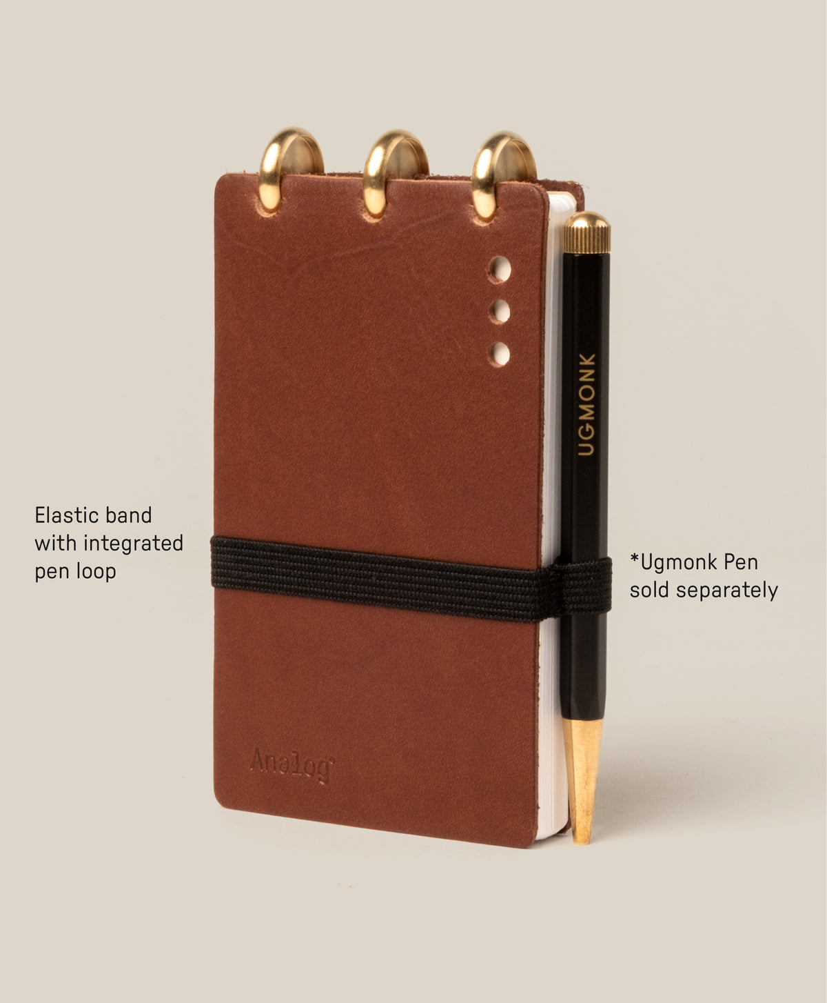 A small brown Leather Discbound Analog notebook with full-grain leather covers, gold rings, and a black elastic band with pen loop. A black and gold Ugmonk pen is beside it; the pen is sold separately.