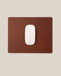 A white Apple Magic Mouse rests on a rectangular Ugmonk Leather Mousepad (Brown) featuring a small embossed logo in the bottom right corner, set against a beige background.