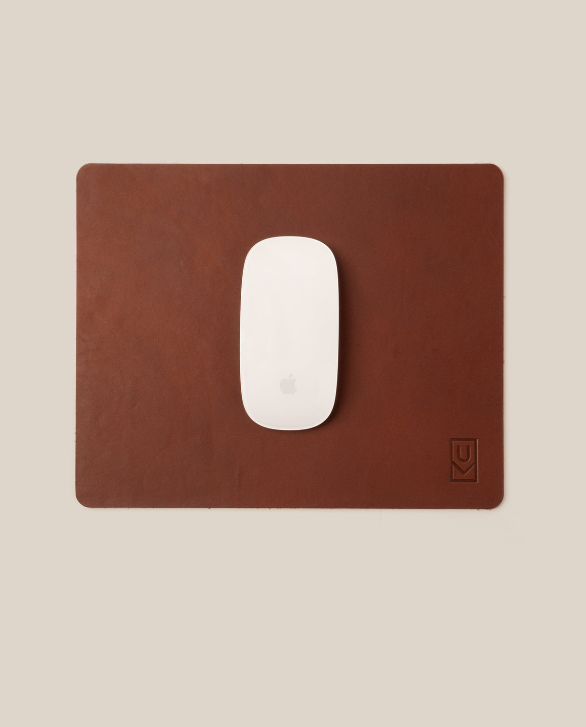 A white Apple Magic Mouse rests on a rectangular Ugmonk Leather Mousepad (Brown) featuring a small embossed logo in the bottom right corner, set against a beige background.