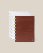 The Discbound Heirloom Journal Bundle (Brown Leather 6" x 8.5" + 3 Refills) is partially open, showing multiple white dot grid pages delicately marked with subtle symbols; crafted from rich brown full-grain leather and accented with elegant gold rings, the refillable discbound journal rests against a plain beige background.