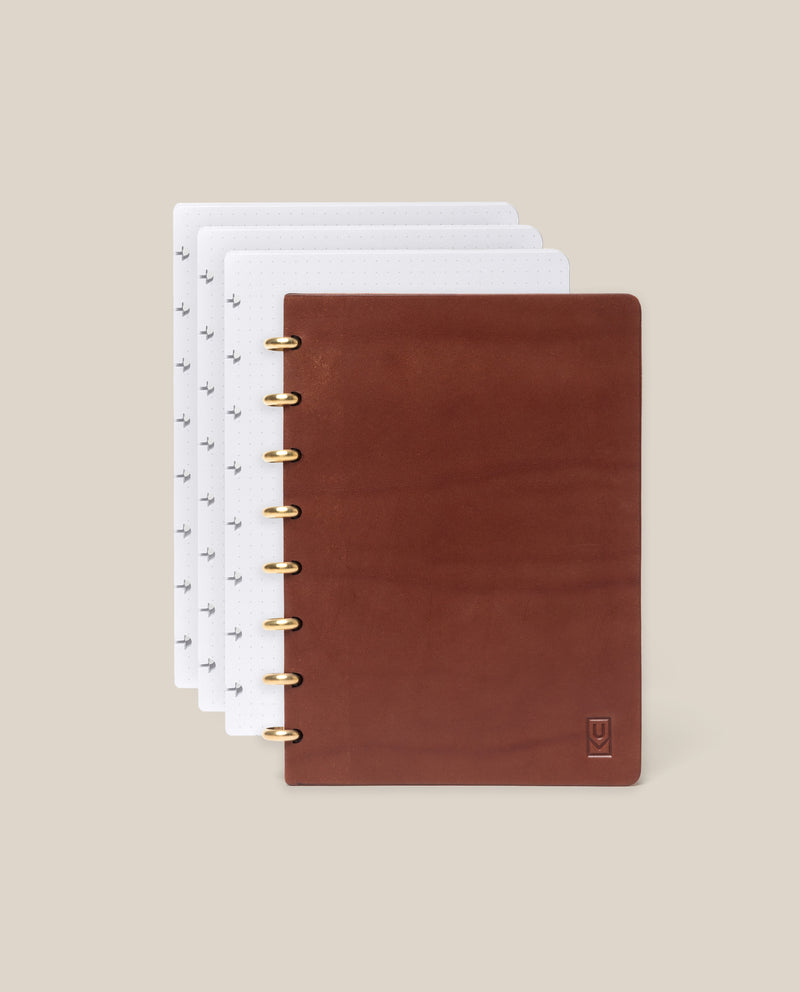 The Discbound Heirloom Journal Bundle (Brown Leather 6" x 8.5" + 3 Refills) is partially open, showing multiple white dot grid pages delicately marked with subtle symbols; crafted from rich brown full-grain leather and accented with elegant gold rings, the refillable discbound journal rests against a plain beige background.
